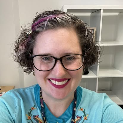 Headshot of Kristin Homuth smiling, with brown hair with purple highlights, black glasses, and a teal shirt
