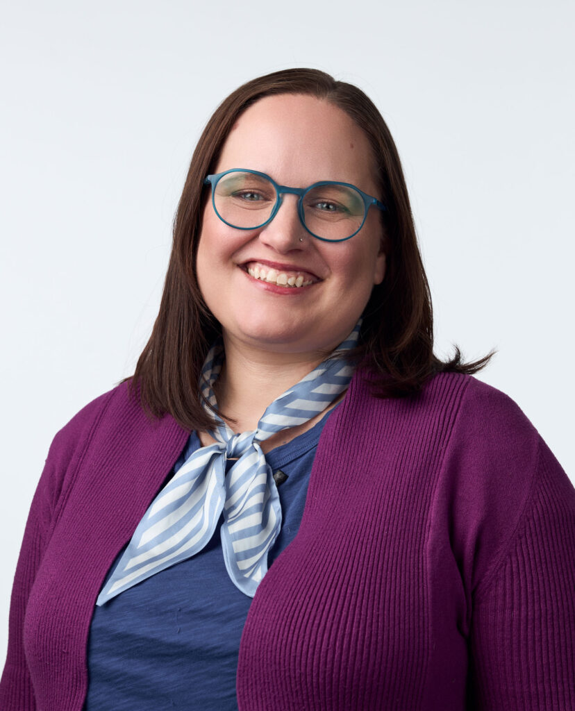 Headshot of Lindsay Tigue smiling, wearing blue glasses, a blue shirt, a purple cardigan, and a patterned scarf